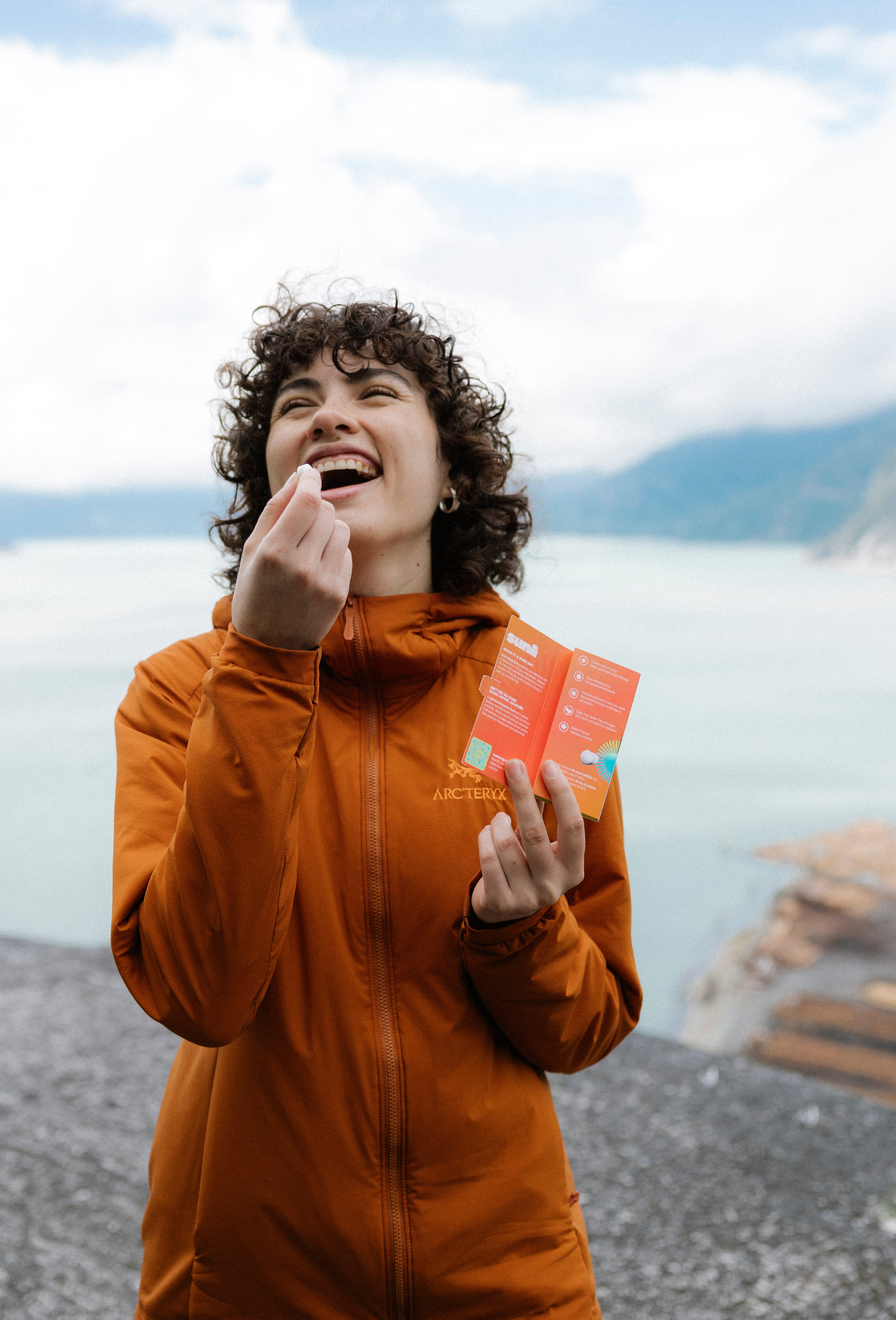 Woman smiling and holding Sunii Energy Gum with a piece of gum in hand, standing by a lakeside